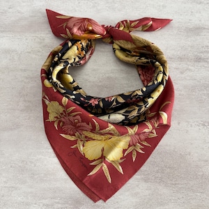 K&ouml;nnte beinhalten: Quadratisches Seidentuch mit floralem Muster in Rot-, Schwarz-, Gelb- und Goldt&ouml;nen. Das Tuch ist oben geknotet und zeigt die leuchtenden Farben und das komplizierte Design. Das Tuch hat eine Signatur in der Ecke.