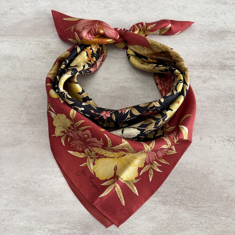 K&ouml;nnte beinhalten: Quadratisches Seidentuch mit floralem Muster in Rot-, Schwarz-, Gelb- und Goldt&ouml;nen. Das Tuch ist oben geknotet und zeigt die leuchtenden Farben und das komplizierte Design. Das Tuch hat eine Signatur in der Ecke.