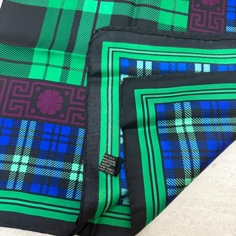 May include: A silk scarf with a plaid pattern in shades of green, blue, and black. The scarf features geometric designs in purple and green. The edges are bordered with a green and black stripe.