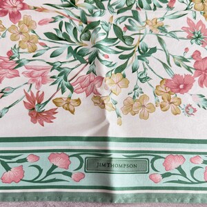 May include: A square scarf featuring a floral pattern with pink, yellow, and green flowers on a white background. The bottom border has a green and light green striped design with pink floral accents and the text JIM THOMPSON in a box.