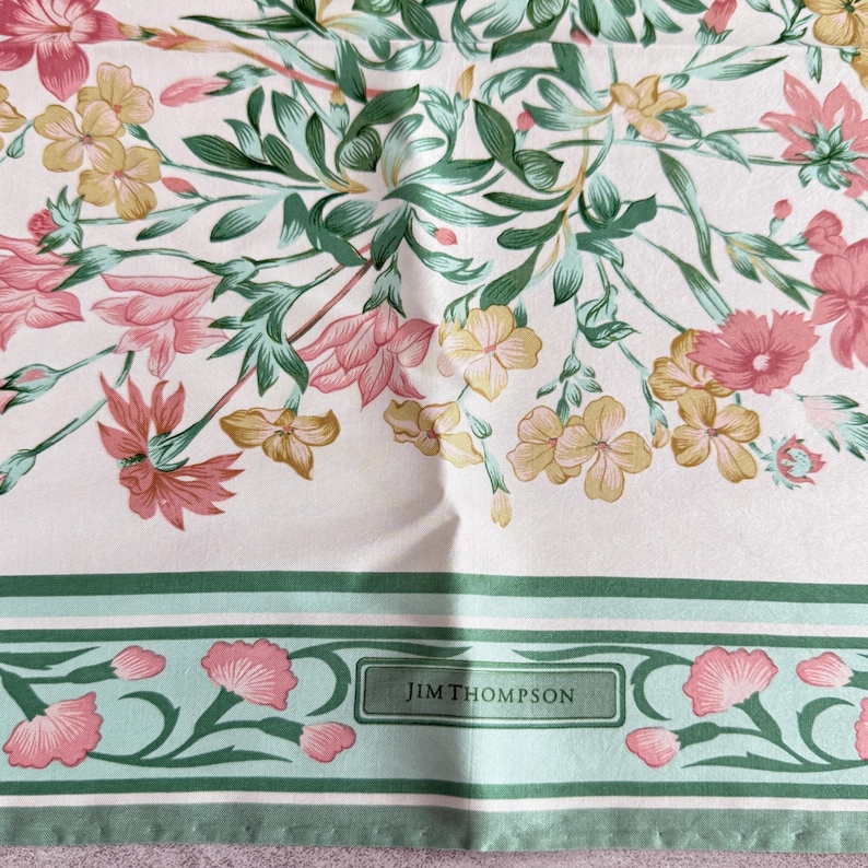May include: A square scarf featuring a floral pattern with pink, yellow, and green flowers on a white background. The bottom border has a green and light green striped design with pink floral accents and the text JIM THOMPSON in a box.