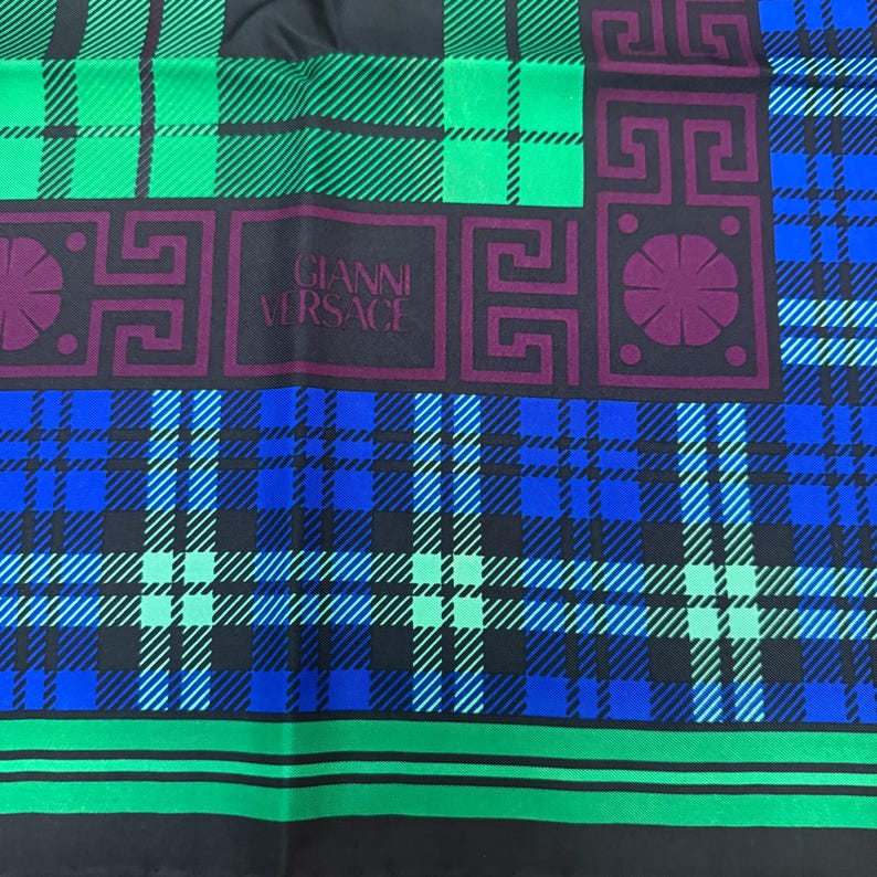 May include: A silk scarf with a plaid pattern in green, blue, and purple. The scarf features the "GIANNI VERSACE" logo in the centre. The scarf has a black border.