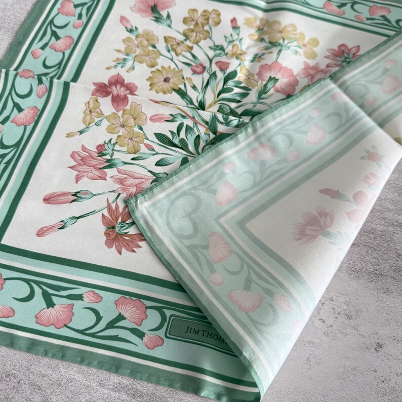 May include: A square silk scarf featuring a white background with a central floral bouquet of pink and yellow flowers. The border is a soft mint green with a repeating floral vine pattern. The text JIM THOMPSON is printed in the bottom corner.