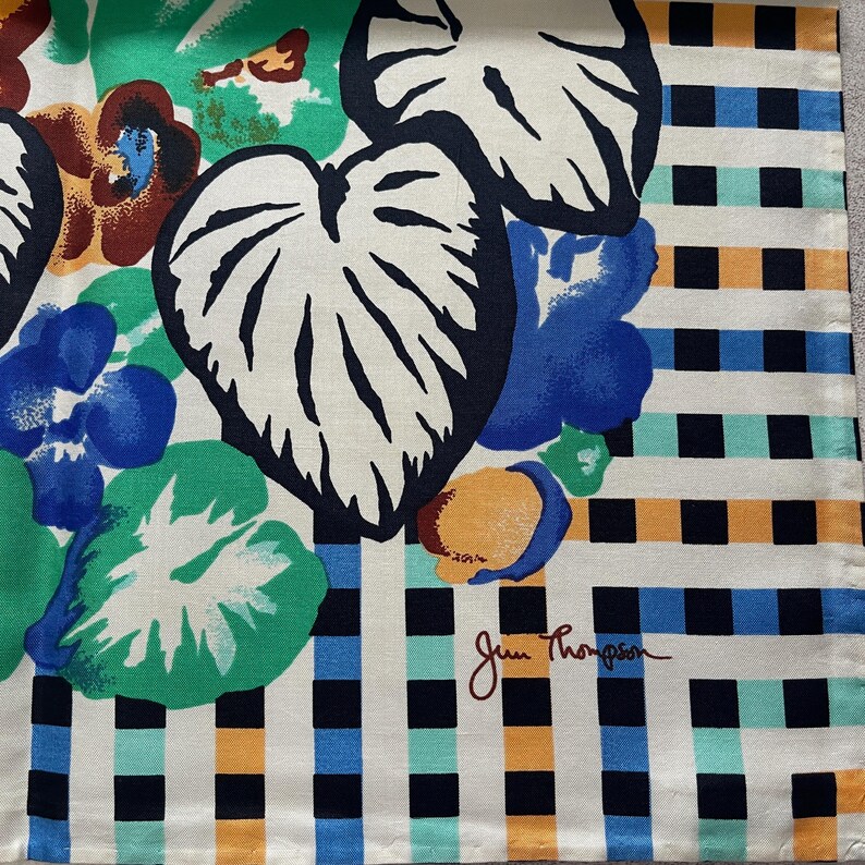 Op de afbeelding: Een witte stof met een geruit patroon in zwart, blauw, groen en oranje. De stof heeft een bloemenmotief met grote bladeren en een handtekening die "Jean Thompson" luidt.