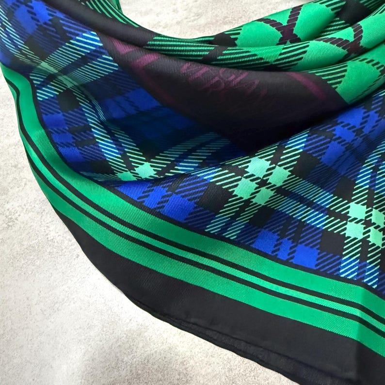 May include: A colourful silk scarf with a plaid design in green, blue, and black. The scarf has a striking green border with black stripes. The fabric is folded, displaying the intricate pattern. The scarf is a stylish accessory.