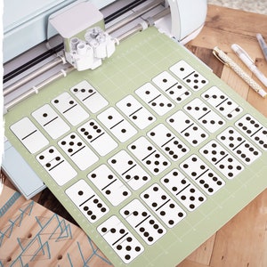 May include: A green cutting mat with a set of 28 black and white dominoes printed on it. The dominoes are arranged in rows and columns. The cutting mat is being used on a Cricut cutting machine.