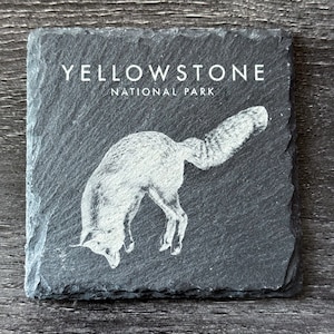 May include: A black slate coaster with a white image of a fox jumping. The text "Yellowstone National Park" is printed in white at the top of the coaster.
