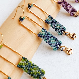 May include: A collection of handmade earrings with gold-colored rectangular frames and woven macrame detailing in shades of green, blue, and purple. Each earring features a small gold-colored lotus charm. The earrings are displayed on a wooden surface.