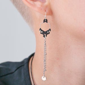 Puede incluir: Un pendiente de plata con un diseño geométrico. El pendiente presenta una forma hexagonal de macramé negro con detalles plateados y una cadena colgante que termina en un pequeño disco. El pendiente se lleva en la oreja.