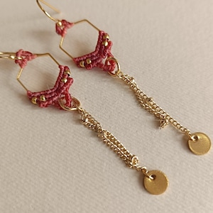 Könnte beinhalten: Ein Paar handgefertigte Ohrringe mit geometrischem Design. Die Ohrringe haben goldfarbene sechseckige Rahmen mit roten Makramee-Details und goldenen Kettenakzenten. Jeder Ohrring hat einen kleinen, runden goldenen Anhänger.
