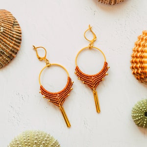 Puede incluir: Un par de pendientes de aro dorados con detalles de macramé marrón y borlas doradas. Los pendientes están fotografiados sobre un fondo blanco con conchas marinas y erizos de mar.