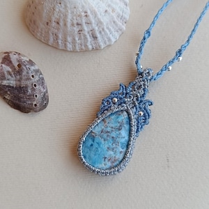 Op de afbeelding: Een handgemaakte macramé ketting met een blauwe stenen hanger. De druppelvormige hanger is gevat in een geweven blauw en zilver macramé ontwerp. Twee schelpen zijn op de achtergrond.