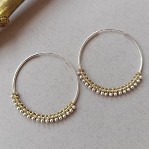 Könnte beinhalten: Ein Paar silberne Creolen mit einem Perlenmuster in Gold und Silber am unteren Rand. Die Ohrringe werden auf einer hellgrauen Oberfläche mit einem goldfarbenen Ast im Hintergrund präsentiert. Ein Schmuckstück.