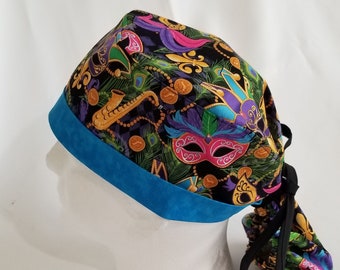May include: A black scrub cap with a vibrant Mardi Gras-themed pattern. The design features masks, coins, and feathers in purple, green, gold, and blue. The cap has a bright blue band and black ties.