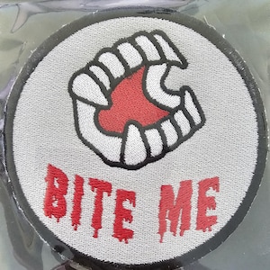 May include: Embroidered patch with a gray background, a black border, and the words "BITE ME" in dripping red letters. The patch features a stylized open mouth with sharp white teeth and a red interior.