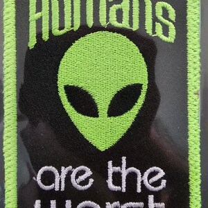 May include: Black patch with a green alien head and the words "Humans are the worst" in white and green. The patch has a green border and is rectangular in shape.