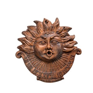 May include: A terracotta sun face with a detailed design, including Roman numerals and the word "MERIDIANA". The sun face has a wide mouth and a prominent nose.