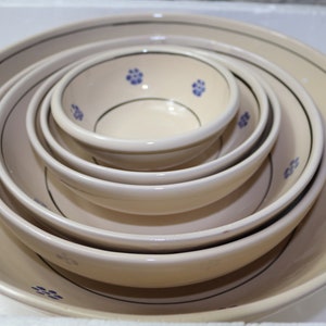 Peut inclure: Ensemble de cinq bols en céramique blanc cassé de tailles différentes, emboîtés les uns dans les autres. Chaque bol est orné d'un bord noir et de petits motifs floraux bleus. Les bols sont disposés du plus grand au plus petit.