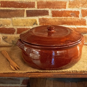 Puede incluir: Una olla grande de cerámica marrón con tapa. La olla tiene un acabado brillante y está sentada sobre una superficie de arpillera.