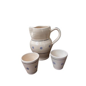 Può includere: Una brocca in ceramica color crema con manico curvo e una coppia di tazze coniche abbinate. Ogni pezzo presenta sottili linee orizzontali nere e piccoli motivi floreali blu dipinti sulle superfici esterne.