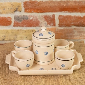 May include: A set of four white ceramic mugs with blue floral designs, a matching ceramic jar with a lid, and a white ceramic tray. The set is on a hessian surface.