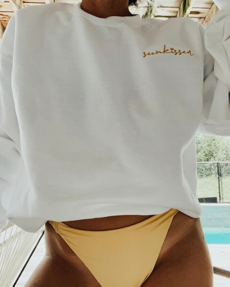 May include: White sweatshirt with the text "sun kisser" embroidered in gold on the front. The sweatshirt is worn by a person wearing a yellow bikini bottom.