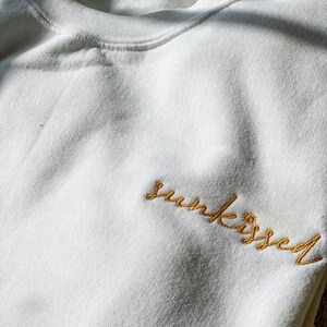 May include: White sweatshirt with the word "sun-kissed" embroidered in gold thread.
