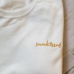 May include: White sweatshirt with the word "sunkissed" embroidered in yellow.