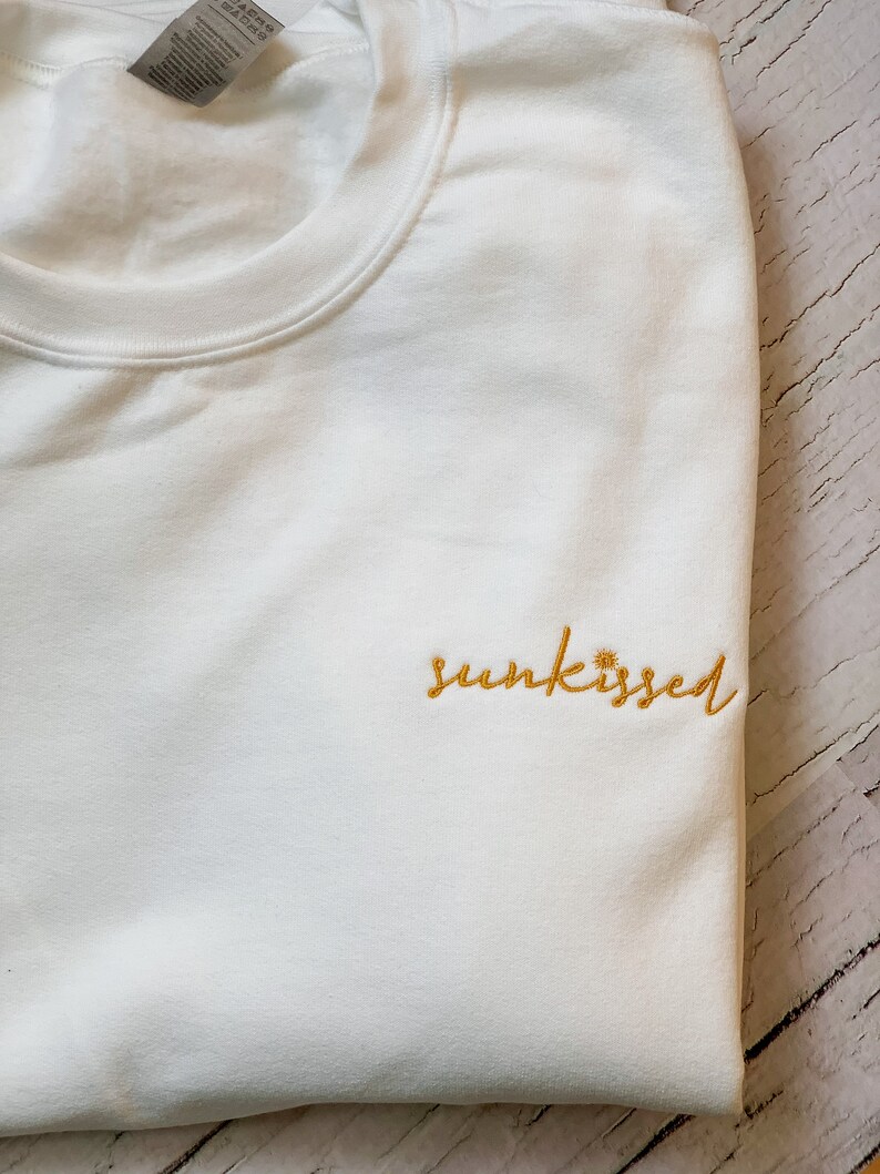 May include: White sweatshirt with the word "sunkissed" embroidered in yellow.