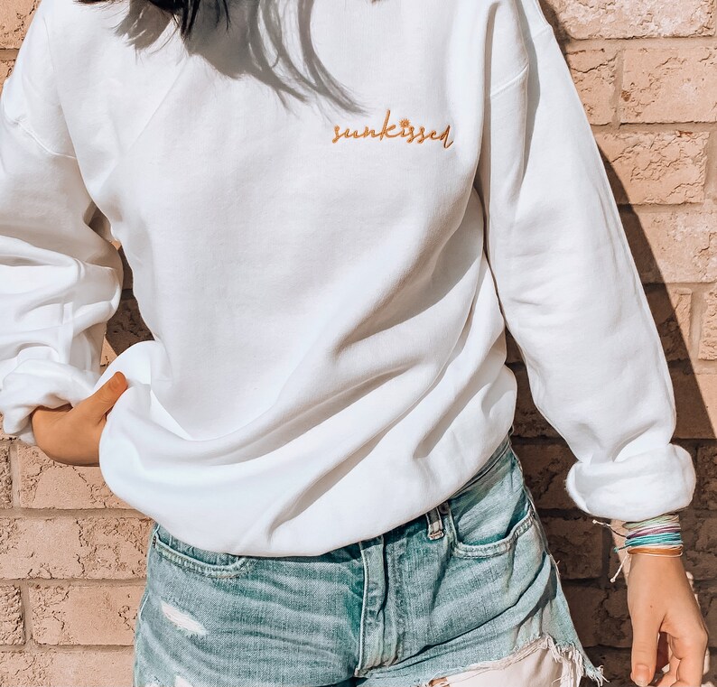 May include: White sweatshirt with the text "sunkissed" embroidered in orange on the front. The sweatshirt is being worn by a person wearing denim shorts.