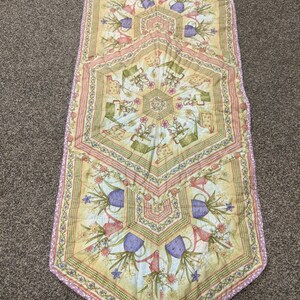May include: A yellow, green, and purple table runner with a geometric pattern of watering cans and flowers. The runner has a scalloped edge and is made of quilted fabric.