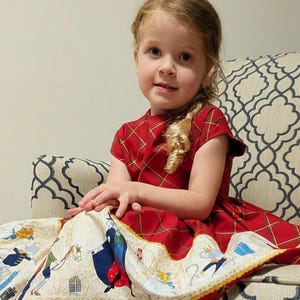 May include: A red dress with a plaid pattern on the bodice and short sleeves. The skirt features a wide cream-colored border printed with illustrations of mice in royal attire, gifts, and horses, finished with a scalloped yellow trim at the hem.
