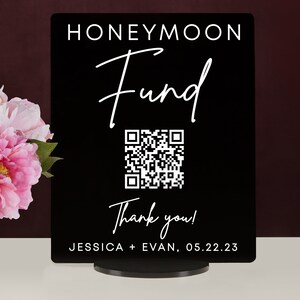 May include: Black sign with white text that reads "Honeymoon Fund" with a stylized cursive font. A QR code is below the text, and below the QR code is the text "Thank you!" and "JESSICA + EVAN, 05.22.23".