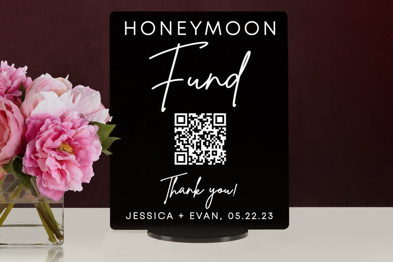 May include: Black sign with white text that reads "Honeymoon Fund" with a stylized cursive font. A QR code is below the text, and below the QR code is the text "Thank you!" and "JESSICA + EVAN, 05.22.23".