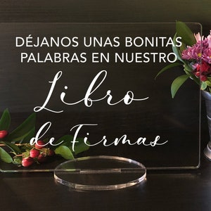 May include: Clear acrylic sign with white script that reads "DÉJANOS UNAS BONITAS PALABRAS EN NUESTRO Libro de Firmas." The sign is displayed on a clear stand, with flowers in a silver vase in the background.