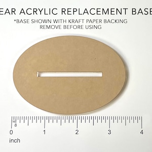 May include: A clear acrylic replacement base for a craft project. The base is oval shaped with a slot cut out of the center. The base is shown with a brown paper backing, which should be removed before using. The base is approximately 3 inches wide.