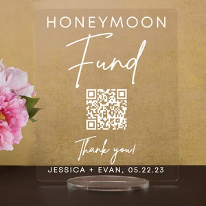 May include: A clear acrylic sign with the text "Honeymoon Fund" in white script font. A QR code is below the text, and the text "Thank you!" is below the QR code. The text "Jessica + Evan, 05.22.23" is at the bottom of the sign.