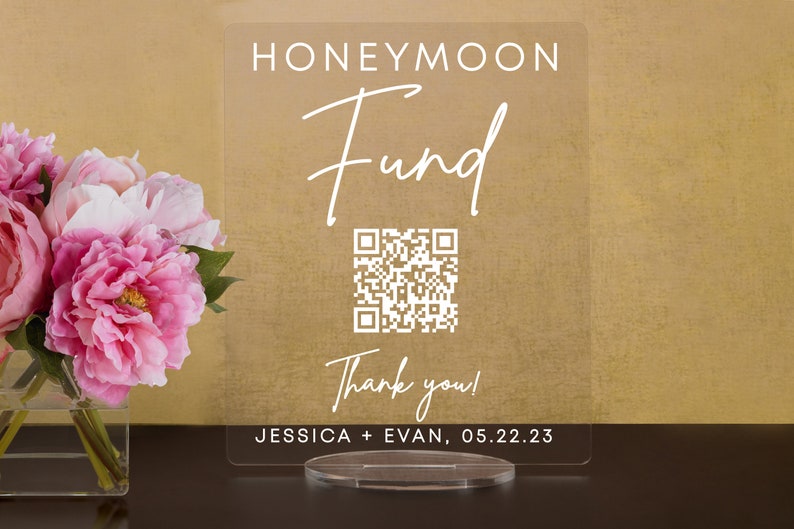 May include: A clear acrylic sign with the text "Honeymoon Fund" in white script font. A QR code is below the text, and the text "Thank you!" is below the QR code. The text "Jessica + Evan, 05.22.23" is at the bottom of the sign.