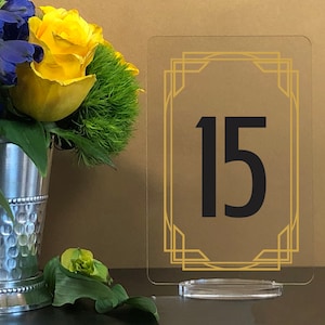 May include: A clear acrylic table number holder with a gold art deco frame and the number 15 in black.
