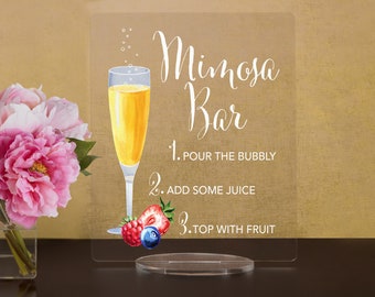 May include: A clear acrylic sign with the text "Mimosa Bar" and instructions for making a mimosa: 1. Pour the bubbly, 2. Add some juice, 3. Top with fruit. The sign features a glass of mimosa with a straw and a few berries at the bottom.