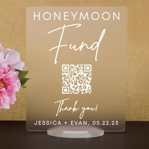 May include: A clear acrylic sign with a gold gradient background. The sign reads "Honeymoon Fund" in white cursive text. A QR code is displayed below the text, followed by "Thank you!" and "JESSICA + EVAN, 05.22.23".