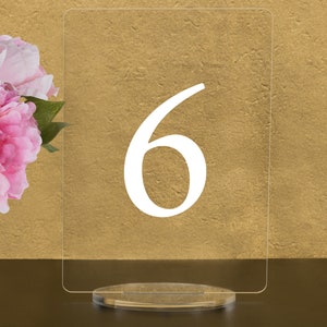 May include: A clear acrylic table number holder with the number 6 printed in white on the front. The holder is on a clear acrylic base.