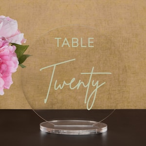 May include: A clear acrylic table number sign with a white base. The sign says "TABLE TWENTY" in a light green font.