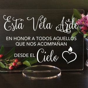 May include: A clear acrylic sign with white text that reads "Esta Vela Arde En Honor A Todos Aquellos Que Nos Acompañan Desde El Cielo" with a white heart and flame graphic.