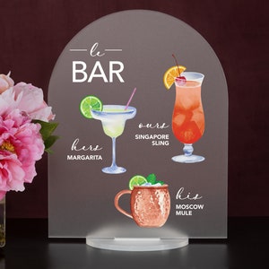 May include: A gray acrylic sign with the text "—le— BAR" and illustrations of three cocktails: a margarita, a Singapore Sling, and a Moscow Mule. The text below each cocktail reads "hers MARGARITA", "ours SINGAPORE SLING", and "his MOSCOW MULE".