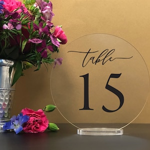 May include: Clear acrylic round table number with the text "Table 15" in black lettering. The table number is on a clear acrylic stand.