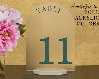 May include: A gold table number card with the number 11 in teal green. The card is shaped like an arch and has the word "TABLE" at the top. The text "Available in FOUR ACRYLIC COLORS" is written in black on the right side of the card.