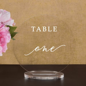May include: Clear acrylic table number with the text "TABLE one" in white lettering. The number is on a clear acrylic stand.