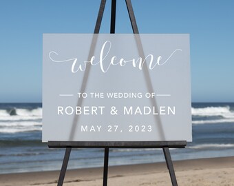 May include: A frosted acrylic wedding welcome sign with the text "Welcome to the wedding of Robert & Madlen May 27, 2023" on an easel.