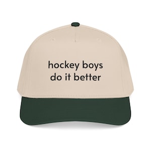 May include: A beige baseball cap with a dark green brim and a dark green button on top. The front of the cap features the black embroidered text "hockey boys do it better" in a simple sans-serif font, centered across the crown of the hat.